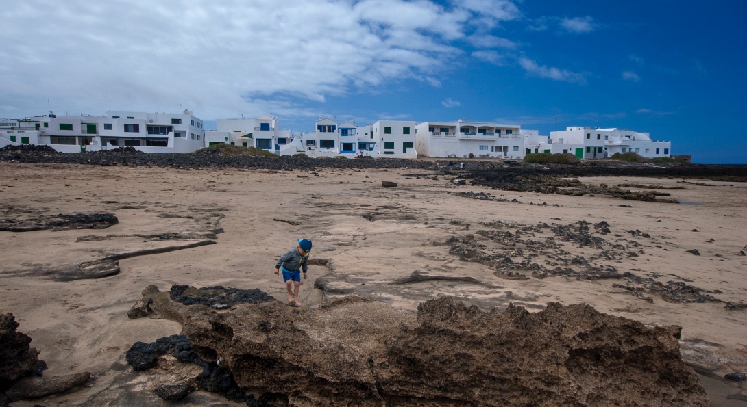 A child explores a rocky, muddy area in front of a group of buildings.